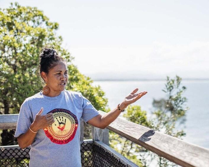 A person standing on a wooden deck with a railing, gesturing with their hands, overlooking a body of water and greenery. 1