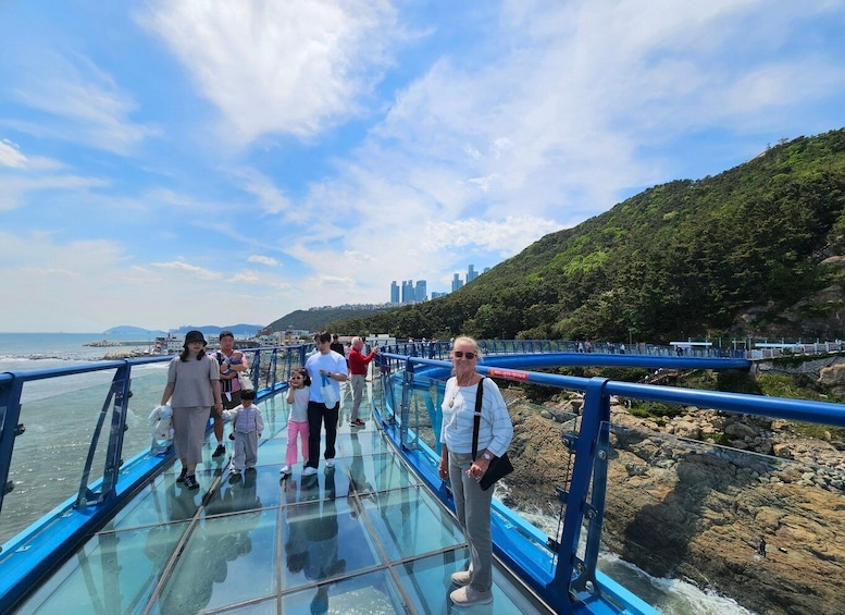 A group of people walking on a glass bridge over water, with a cityscape and hills in the background. 5