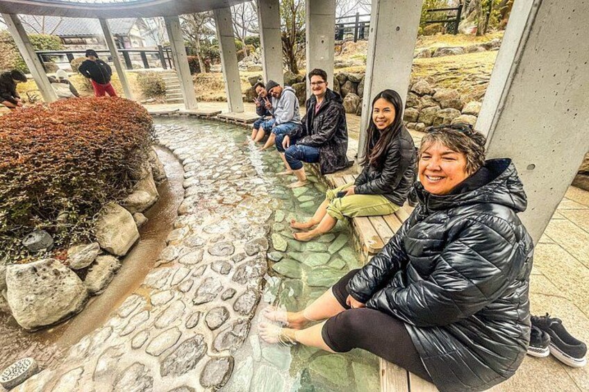 A group of people sitting on benches under a shelter, with a stone pathway and a small water feature in the background. 1
