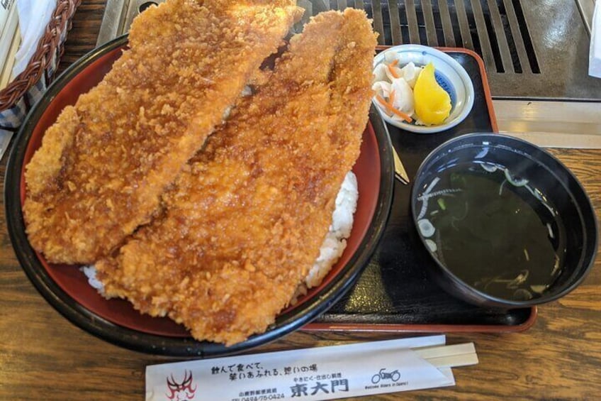 A plate of breaded and fried fish served over rice, accompanied by a small bowl of dipping sauce and a side of pickled vegetables. 4