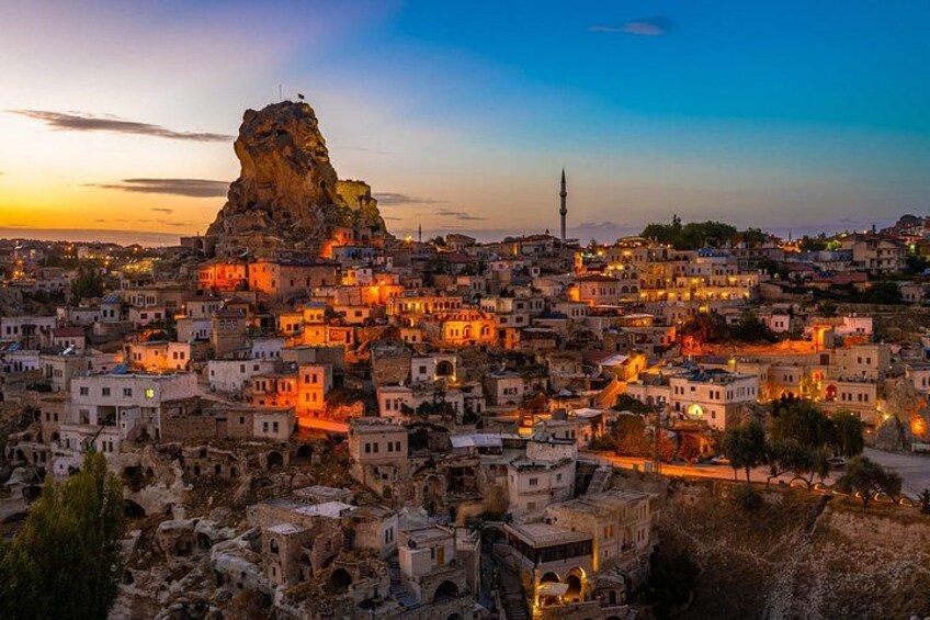 Une vue sur une ville au crépuscule, avec des bâtiments illuminés, une formation rocheuse imposante et un minaret visible au loin. 4