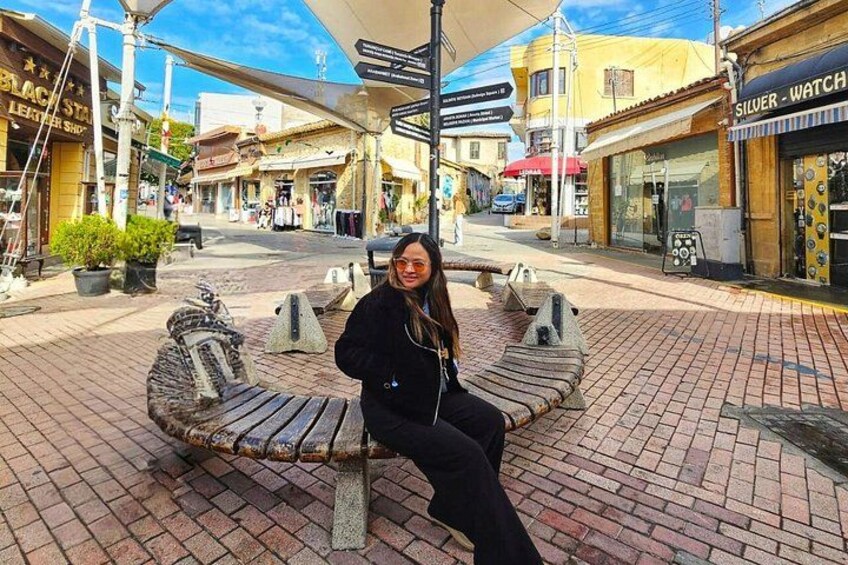 Una mujer sentada en un banco en una zona peatonal con tiendas y una farola. 2