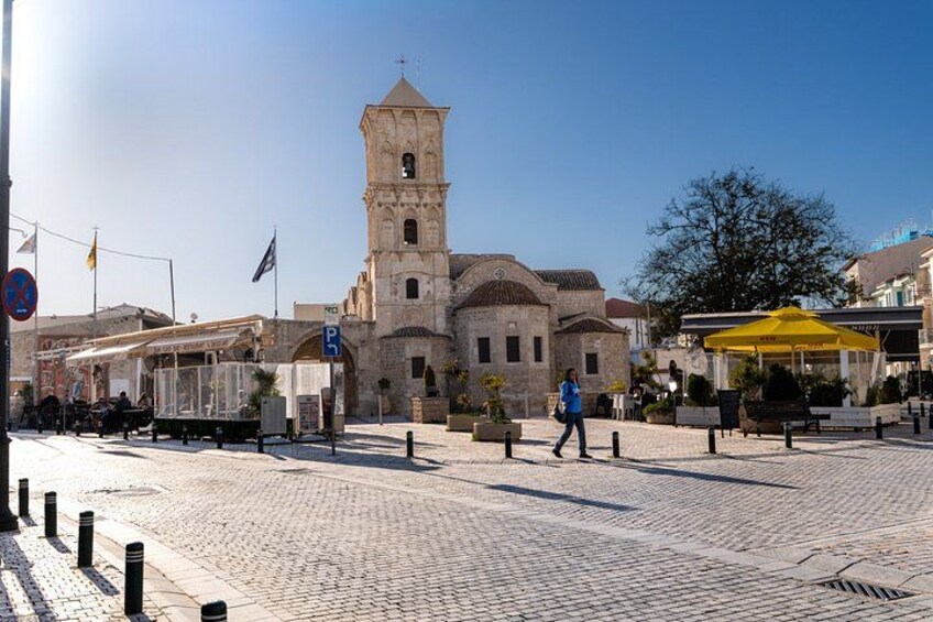 A cobblestone square with a historic building featuring a tower, a café with a yellow umbrella, and a pedestrian walking by. 1