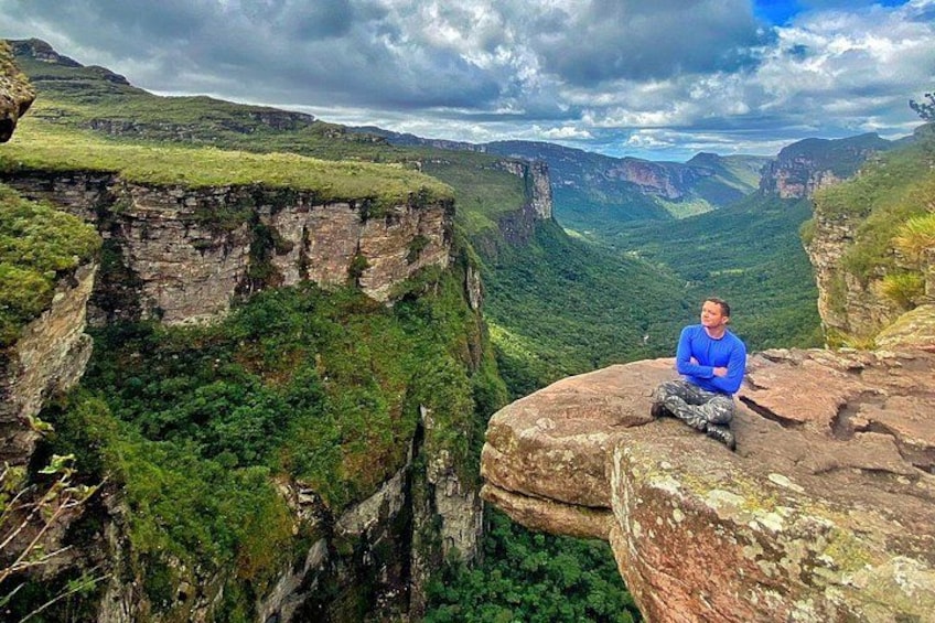 A person sitting on a rock ledge overlooking a deep valley with steep cliffs and dense greenery. 5
