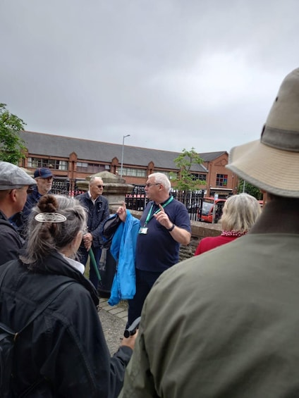 A group of people gathered outdoors, some wearing jackets and hats, in front of a building with a brick facade and a sign that reads "Sandhurst." 1