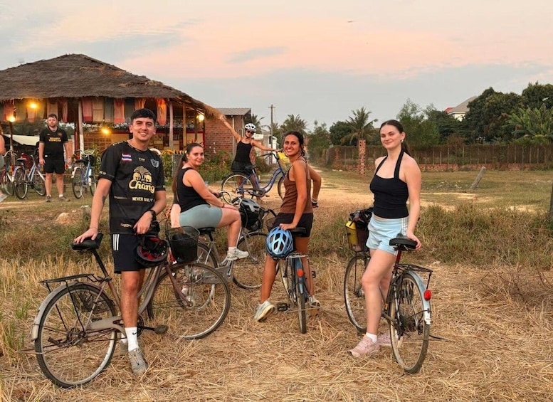 A group of people on bicycles in an outdoor setting with a thatched-roof building and trees in the background. 4