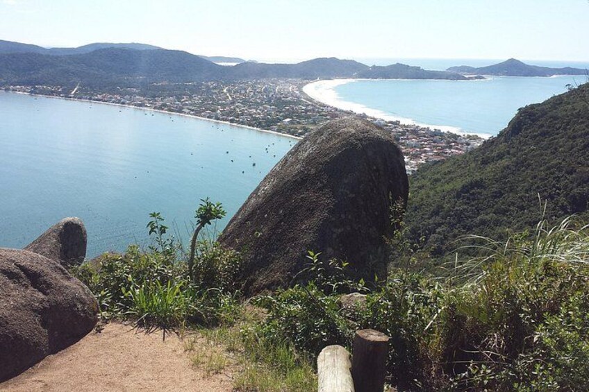 A coastal landscape with a large rock formation, a sandy path, and a view of a bay with a town and mountains in the distance. 3