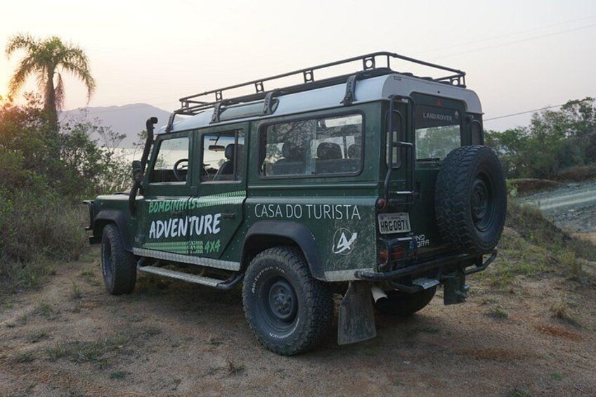 A green off-road vehicle with 'BOMBENHITS ADVENTURE' and 'CASA DO TURISTA' written on it, parked on a dirt path with a palm tree and mountains in the background. 4