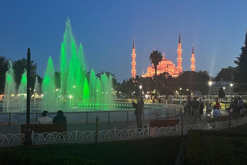 A fountain with green lights, a mosque with illuminated minarets, and people in a park at dusk. 4