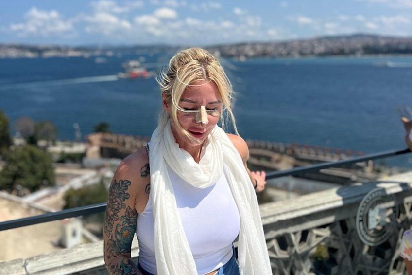 A person with tattoos on their arm, wearing a white tank top and a white scarf, standing on a bridge with a view of a body of water and distant land. 3