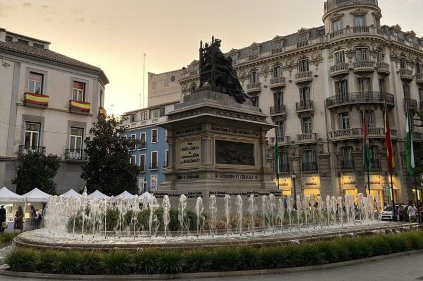 A fountain with water jets, surrounded by a circular walkway, in front of a historic building with balconies and flags. 3