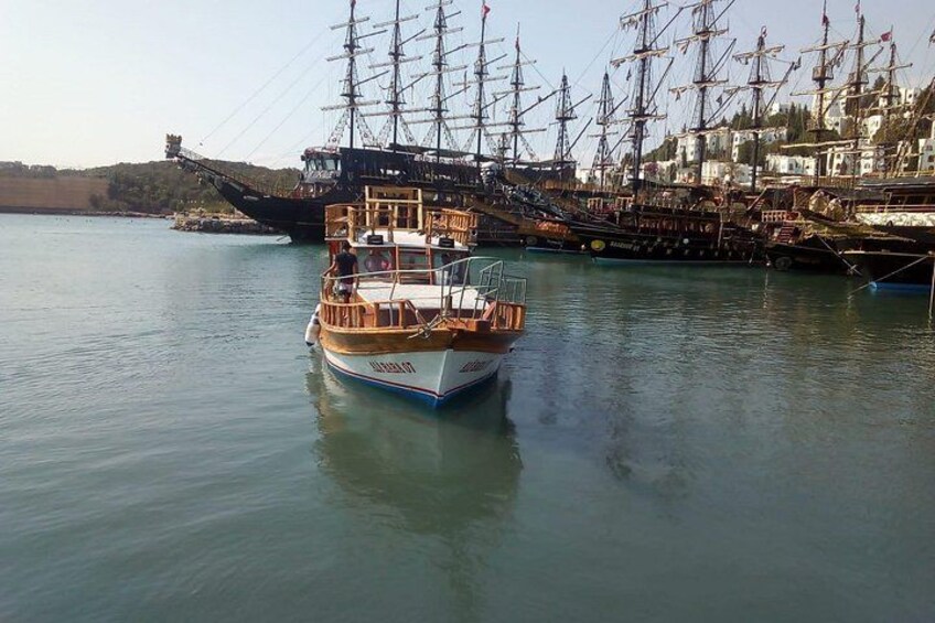 Een klein bootje met een houten dek en een witte romp ligt afgemeerd nabij een groter schip met meerdere masten. 2