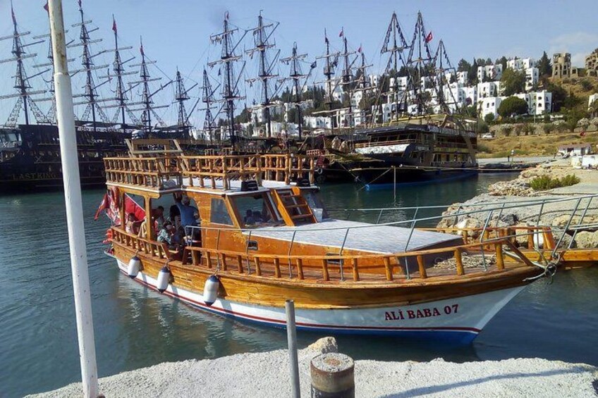 Een houten boot met de naam 'ALI BABA 07' afgemeerd in een jachthaven, met grote schepen op de achtergrond. 1