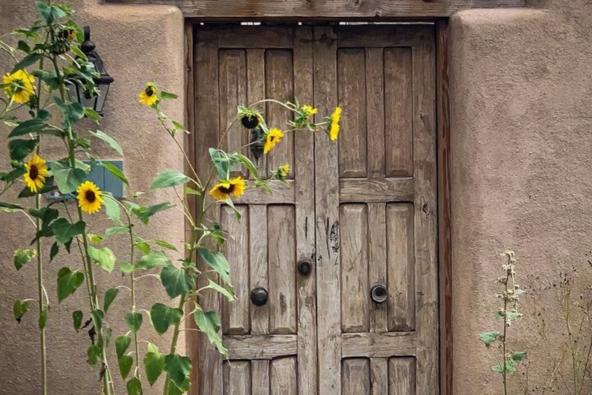 A rustic wooden door with a weathered texture, surrounded by a stucco wall and a plant with yellow flowers. 1