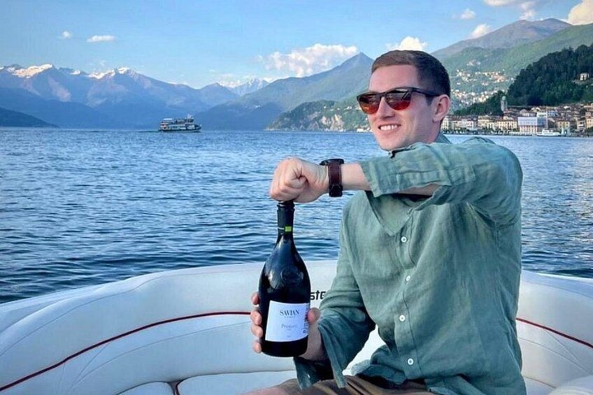 En person holder en vinflaske på en båd med en sø og bjerge i baggrunden. 4