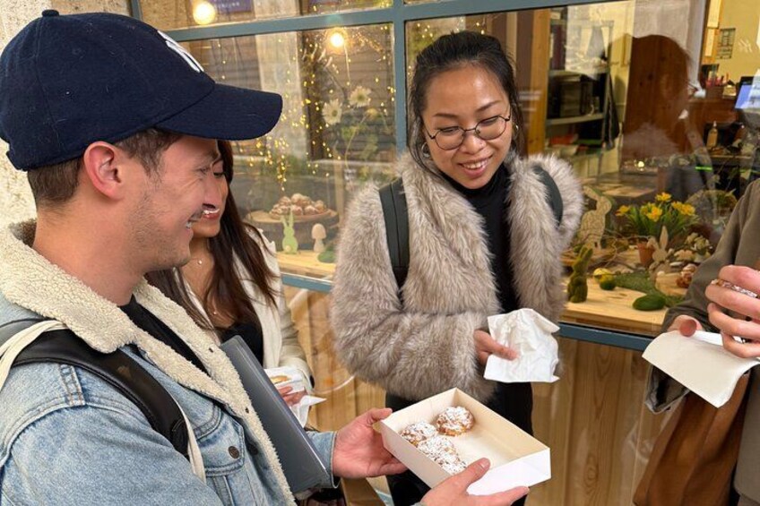 En person holder en æske med donuts, en anden person er iført en pelsfrakke, og et butiksvindue prydes af blomster. 2