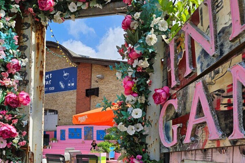Eine lebhafte Straßenszene in der Stadt mit einem pink-weißen Blumengitterbogen, einem Schild mit der Aufschrift 'IN GARDEN' und Graffiti sowie einem blauen Schild für 'Street Food'. 3