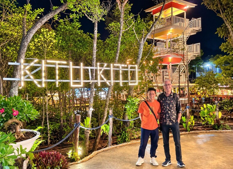 Two people standing in front of a sign with illuminated letters, surrounded by greenery and a building with a red roof. 1