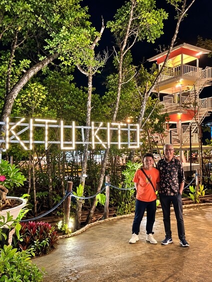 Two people standing in front of a sign with illuminated letters, surrounded by trees and a building with a balcony. 2