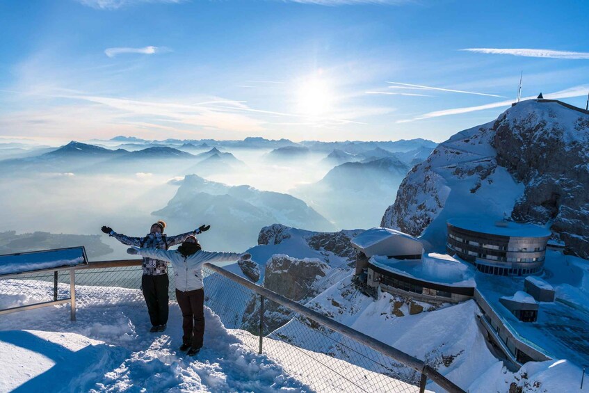 Two people standing on a snowy mountain with a view of distant mountains and a snow-covered building. 3