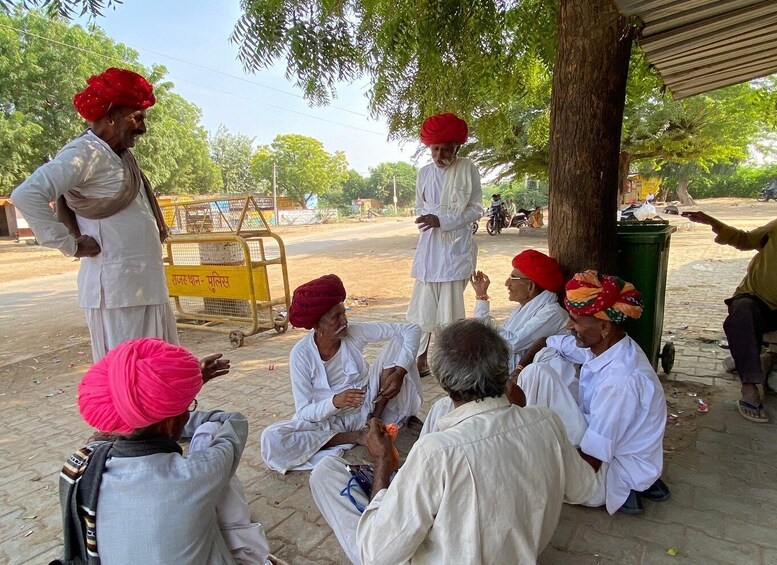 A group of men in traditional attire sitting and standing outdoors, engaged in a discussion. 1