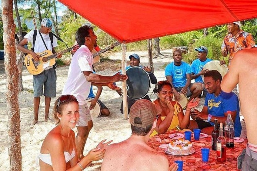 A group of people gathered around a table with food and drinks, under a red canopy, on a sandy beach. 5