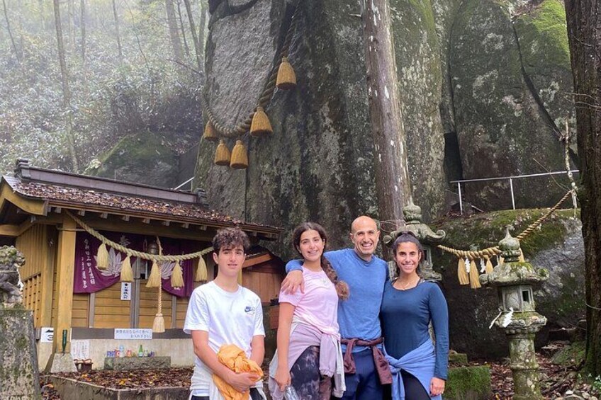 A group of people standing in front of a traditional building with a thatched roof, surrounded by a forested area with large rocks and a rope ladder. 1