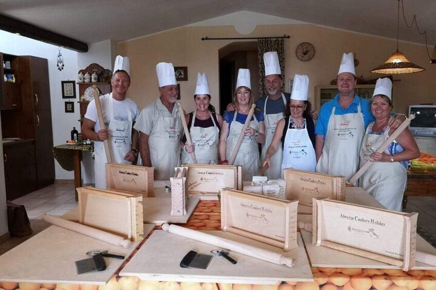 A group of people in chef hats and aprons standing in a kitchen with baking tools and boxes labeled 'Merry Christmas Cookies.' 1