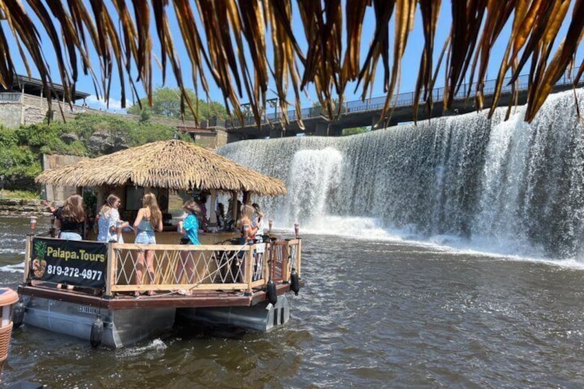 A boat tour with a thatched roof, passengers, and a waterfall. 1