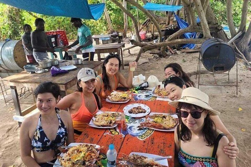 A group of people sitting at an outdoor table with food, under a blue canopy, with trees and a barbecue grill in the background. 5