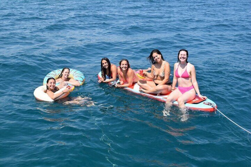 Five people floating on a body of water using inflatable rings and a paddleboard. 5
