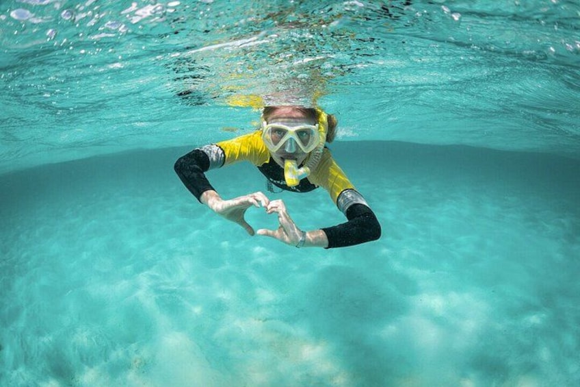 A person underwater, wearing a yellow and black wetsuit, making a heart shape with their hands. 1