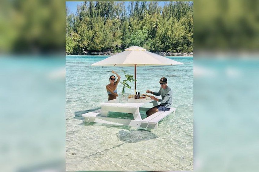 A couple dining at a white table under a large umbrella on a boat in clear tropical waters. 3