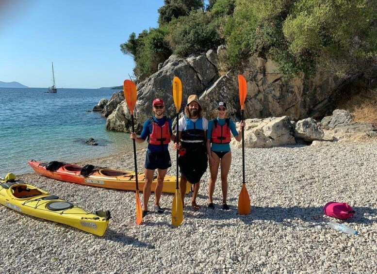 Three people on a pebbled beach with kayaks and paddleboards, a sailboat in the distance, and rocky cliffs. 3