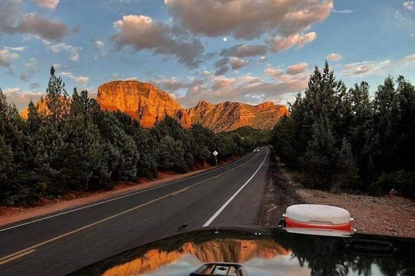 A road with a car's dashboard in the foreground, surrounded by trees and a mountain in the background. 2