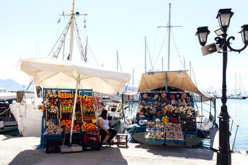 Un mercado frente al mar, con puestos que venden frutas y mariscos, barcos amarrados al fondo y un farol. 2