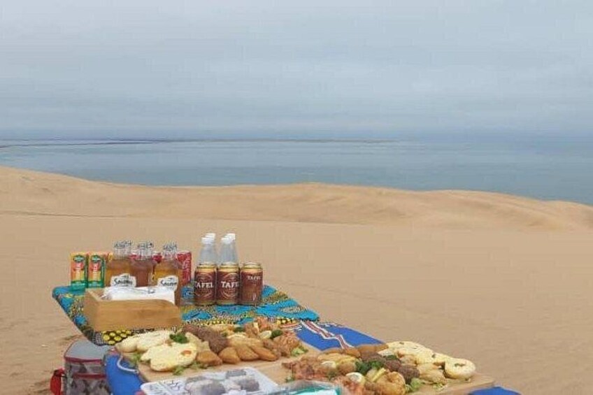 A picnic setup on a sandy beach with food, drinks, and a tablecloth. 4