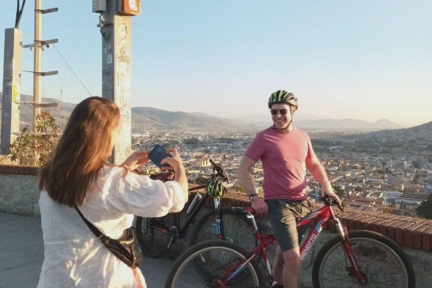 Iemand maakt een foto met een camera, een andere persoon staat naast een fiets, en een stadsgezicht op de achtergrond. 4