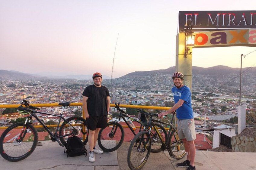 Twee personen staan naast fietsen, met een stadsgezicht en bergen op de achtergrond. 5