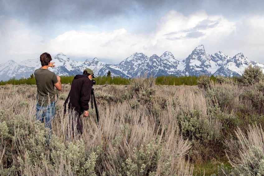 Two people with cameras, a grassy field, and snow-capped mountains in the background. 1