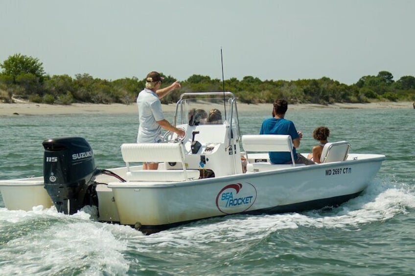 A white boat with the text "Sea Rocket" and "MD 2697 CT" on its side, moving on the water with a sandy beach and greenery in the background. 2