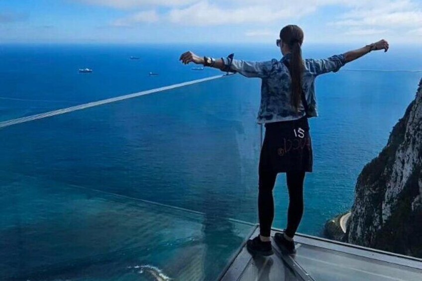 A person standing on a glass bridge with arms outstretched, overlooking a body of water and a distant coastline. 5