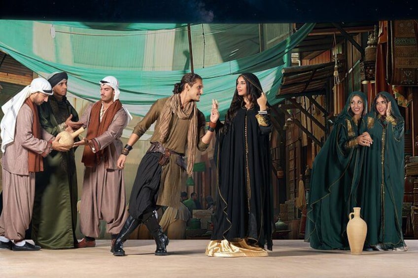 A group of people in historical costumes on a stage with a backdrop of bookshelves and a large vase. 4