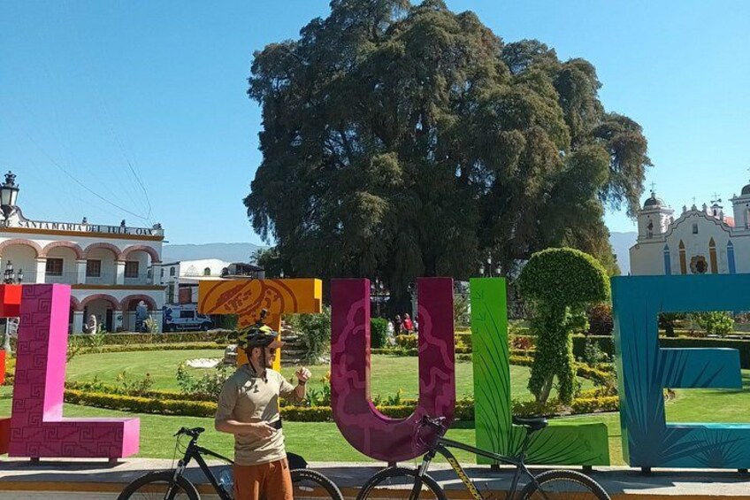Una persona de pie frente a grandes letras coloridas que deletrean 'LULU', con una bicicleta y un edificio al fondo. 1