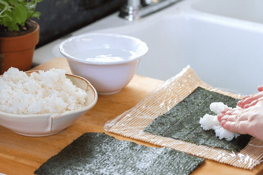 A bowl of white rice, a bowl of white liquid, and a hand placing rice on a seaweed sheet on a wooden board. 2