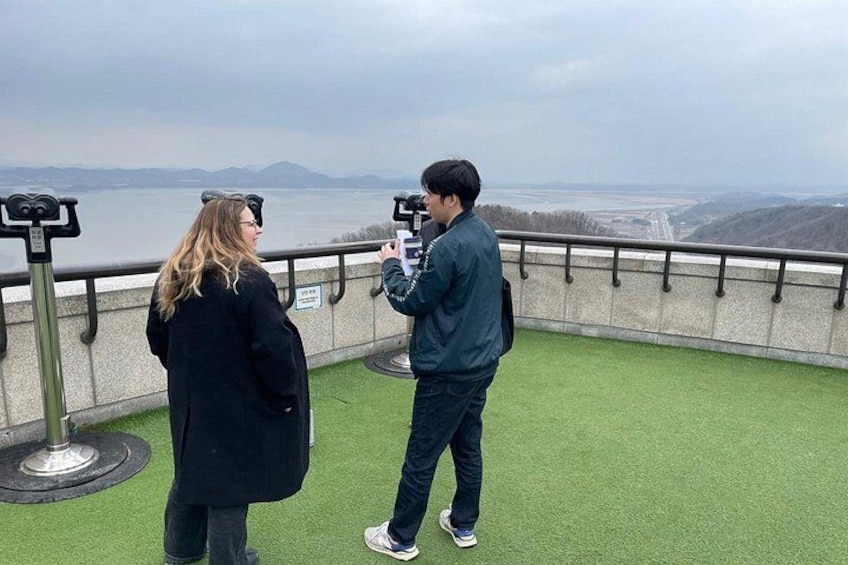 Deux personnes se tiennent sur une plateforme verte entourée d’une rambarde, d’où elles dominent une étendue d’eau et un paysage lointain. 2