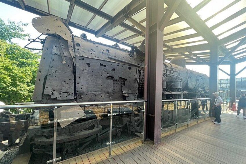 Una locomotora grande y oxidada exhibida en una estructura cubierta con piso de madera y barandilla metálica. 3