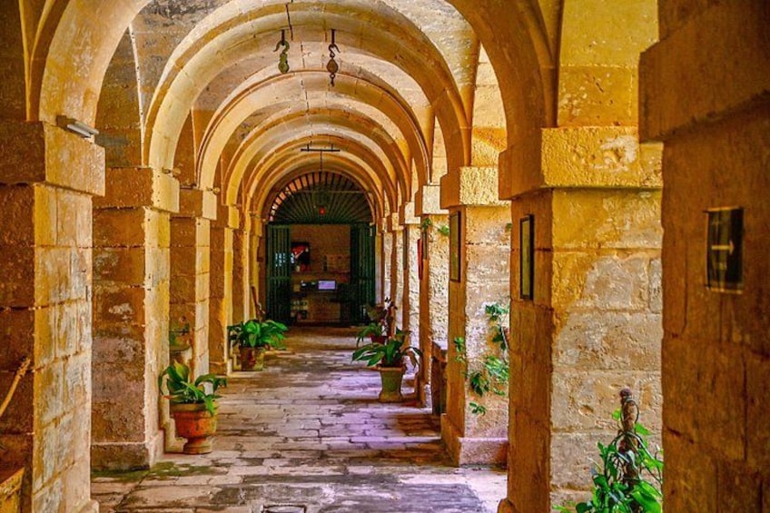 A stone corridor with arches, potted plants, and a view into a room with a green door. 4