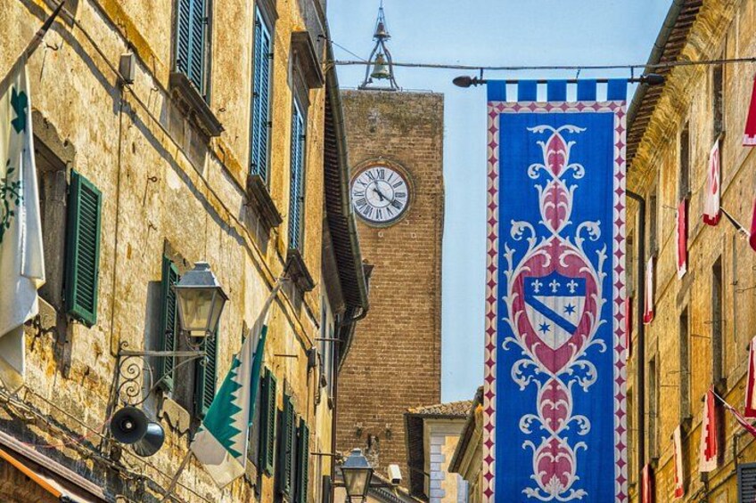 A narrow street with buildings, a clock tower, and a banner with a crest. 2
