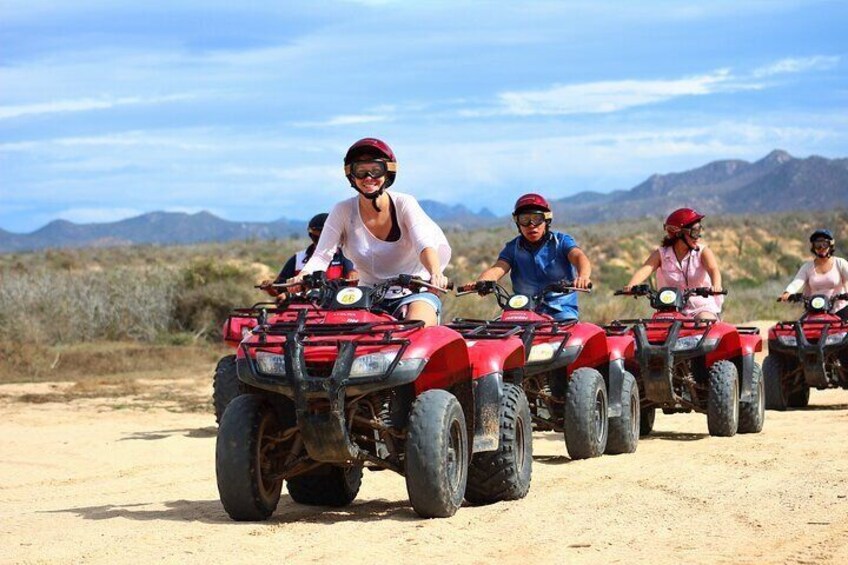 Vier personen rijden op rode quads over een zandweg, met een woestijnlandschap en bergen op de achtergrond. 1
